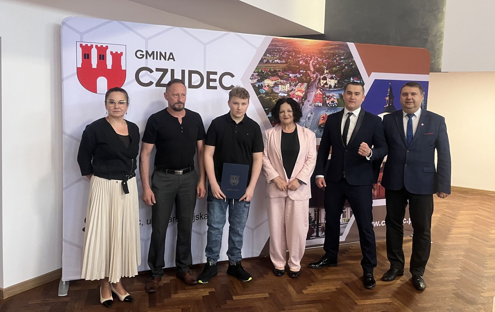 Zdjęcie na tle ścianki promocyjnej nagrodzonego wraz z przedstawicielami Gminy Czudec. 
